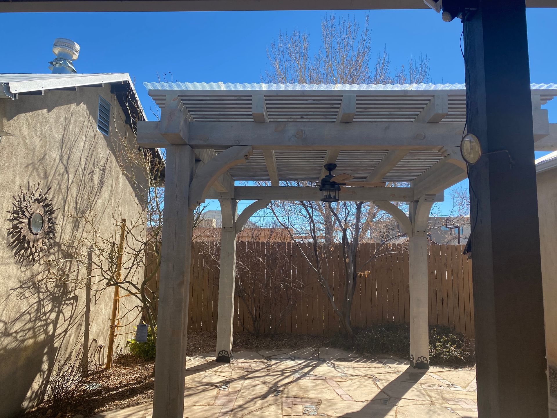 Pergola with fan in a sunlit outdoor area, with a building on the left and a fence in the background.