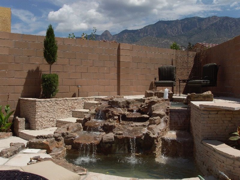 Backyard stone waterfall and pond beside tan block walls, with mountains and cloudy sky in the background