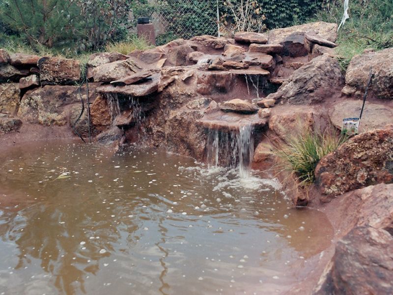 Rock waterfall spilling into a muddy pond with stone ledges and surrounding greenery