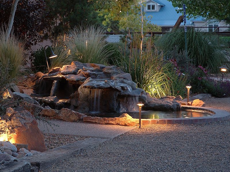 Illuminated backyard rock waterfall and pond beside a gravel path at dusk