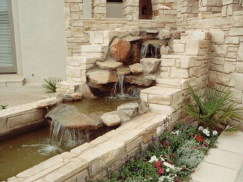 Small stone fountain spilling water into a narrow pond beside a walkway and flower bed