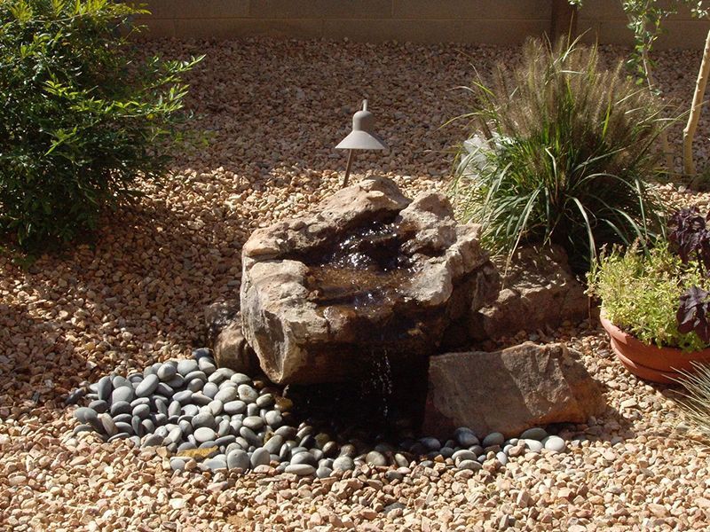 Small garden waterfall fountain surrounded by pebbles and plants in a sunlit yard