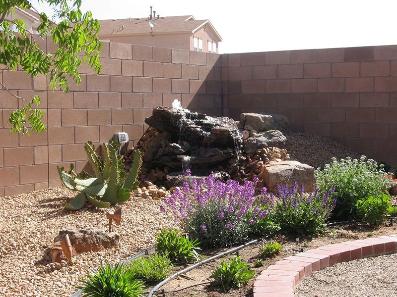 Desert backyard with a rock waterfall, flowering shrubs, cactus, and a curved brick border