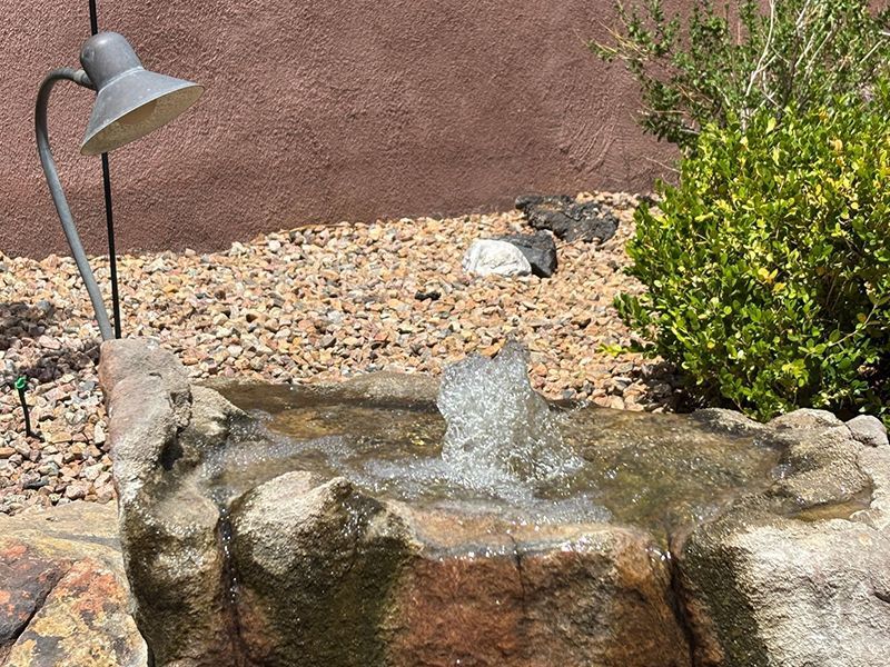 Small backyard stone fountain with running water, a lamp, pebbles, and green shrubbery against a stucco wall