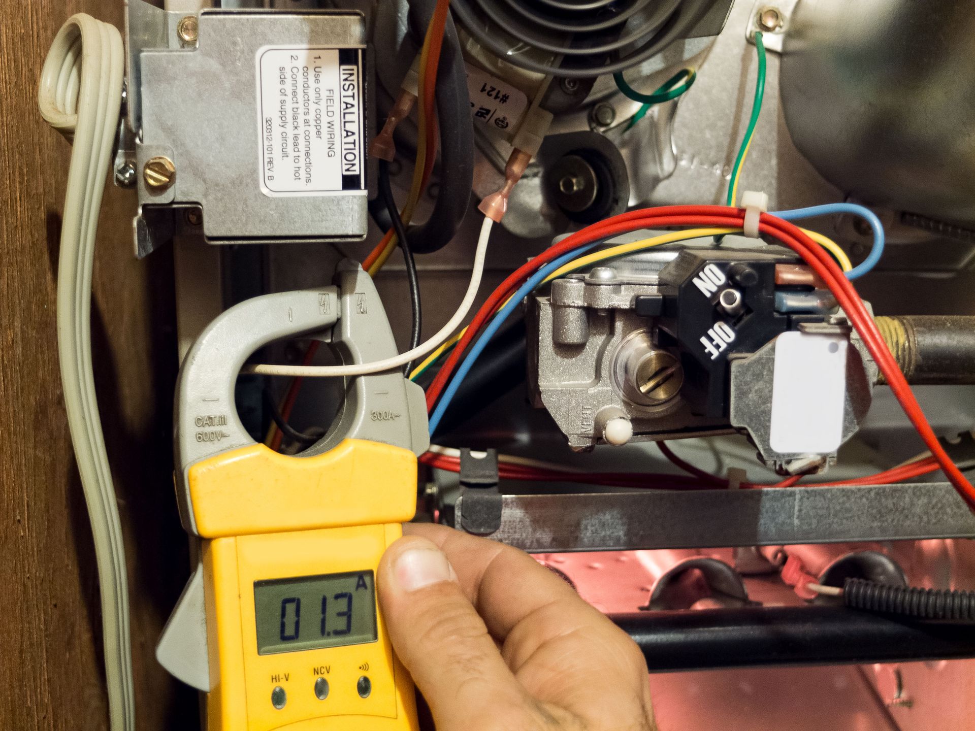 A hand holds a clamp meter on furnace wiring, reading 0.13 amps.