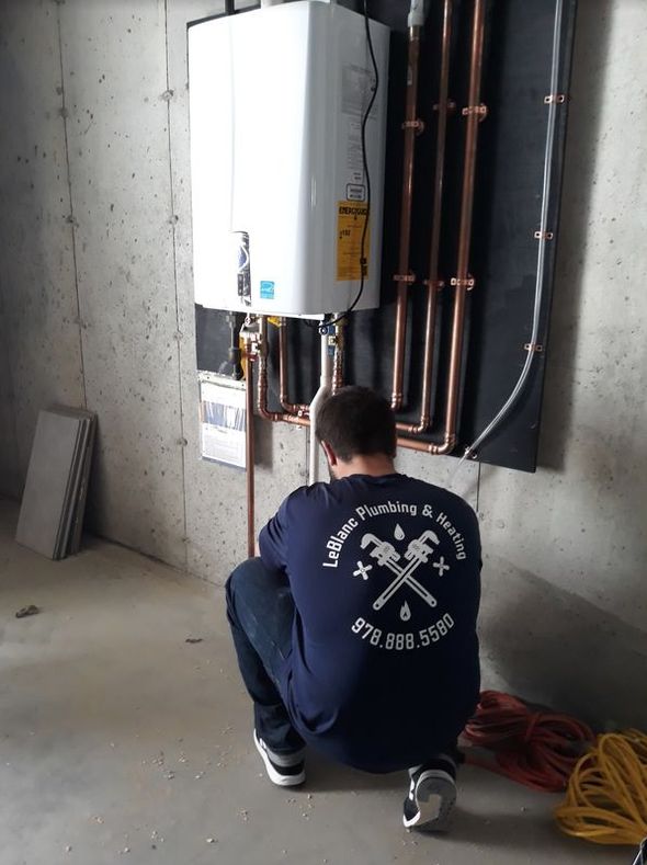 Plumber inspecting a wall-mounted water heater in a concrete-walled basement. Copper pipes and wiring are visible.