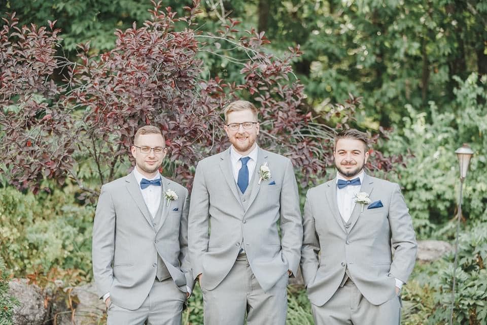 Three men in gray suits pose outdoors in front of greenery.