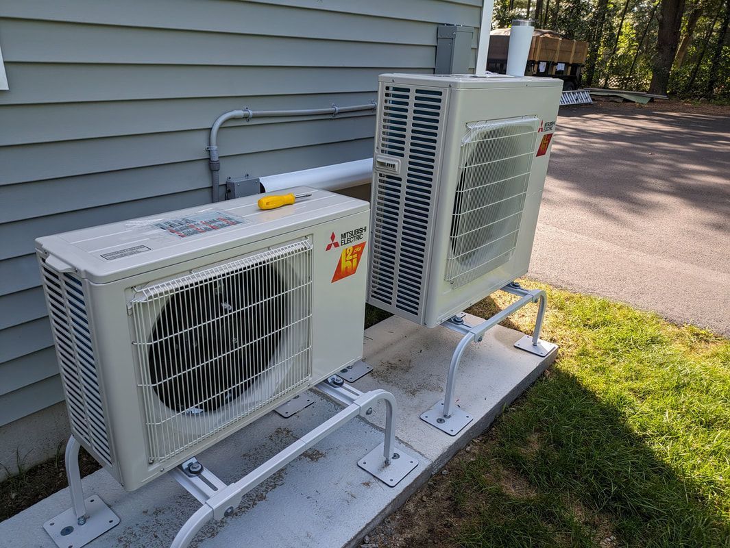 Two beige heat pump units on concrete pad, mounted outside a house on metal brackets.