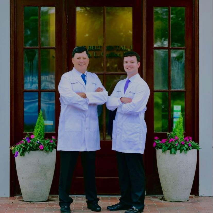 Two men in white coats stand in front of a building with crossed arms.