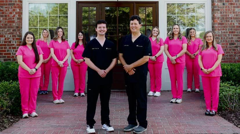 Two men in black scrubs stand before a group of women in pink scrubs, posing in front of a building.