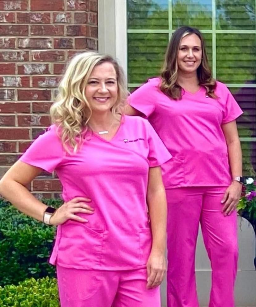 Two women in pink scrubs smiling outside a brick building.