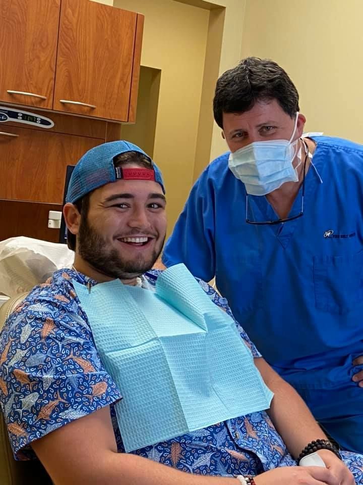 Man in dental chair with a bib, smiling, dentist in scrubs and mask smiles. Dental office setting.