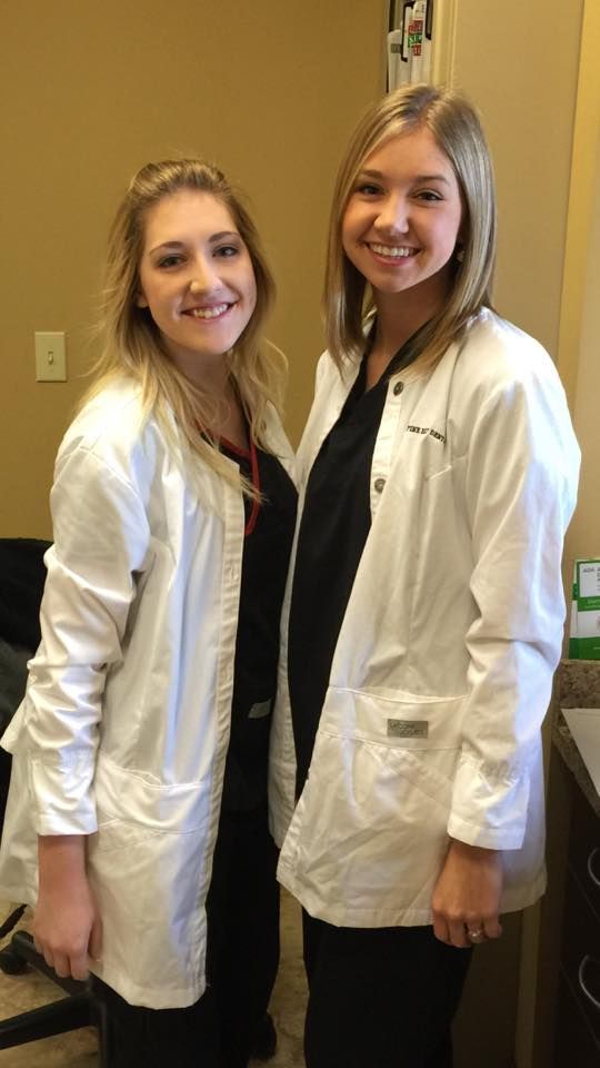 Two women in white lab coats and black scrubs, smiling. Inside a building.