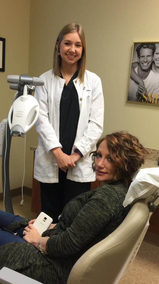 Woman in dental chair, dental assistant in white coat stands behind. Room with whitening equipment.