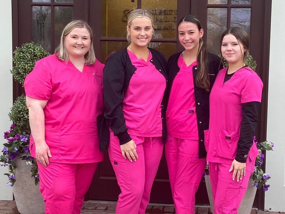 Four people wearing matching bright pink scrubs stand together in front of a doorway with potted plants on either side.
