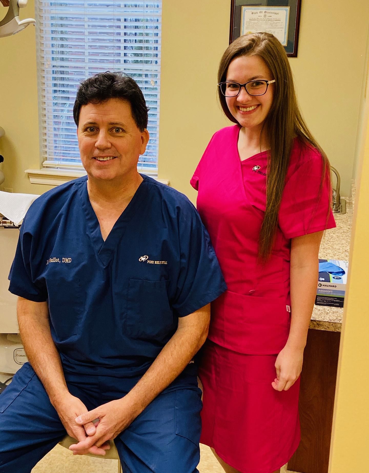 A man in navy blue scrubs and a woman in a pink scrub top and skirt smiling in a professional office setting.