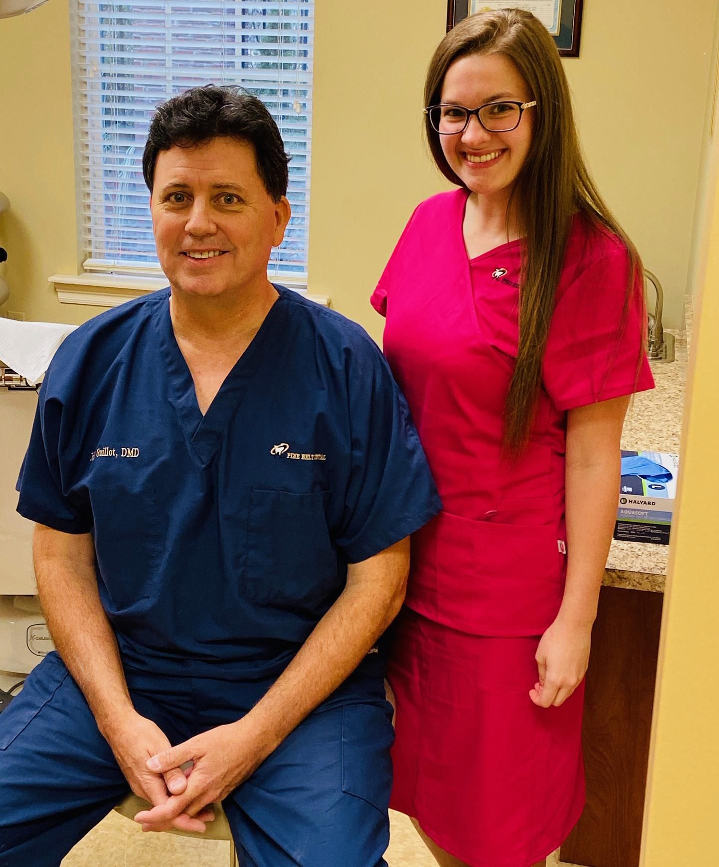 A professional in blue scrubs sits next to a colleague in pink scrubs in an office, both smiling at the camera.