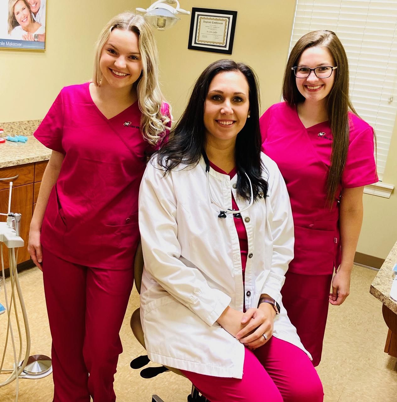Three dental professionals in a clinic, one in a white coat, two in pink scrubs.