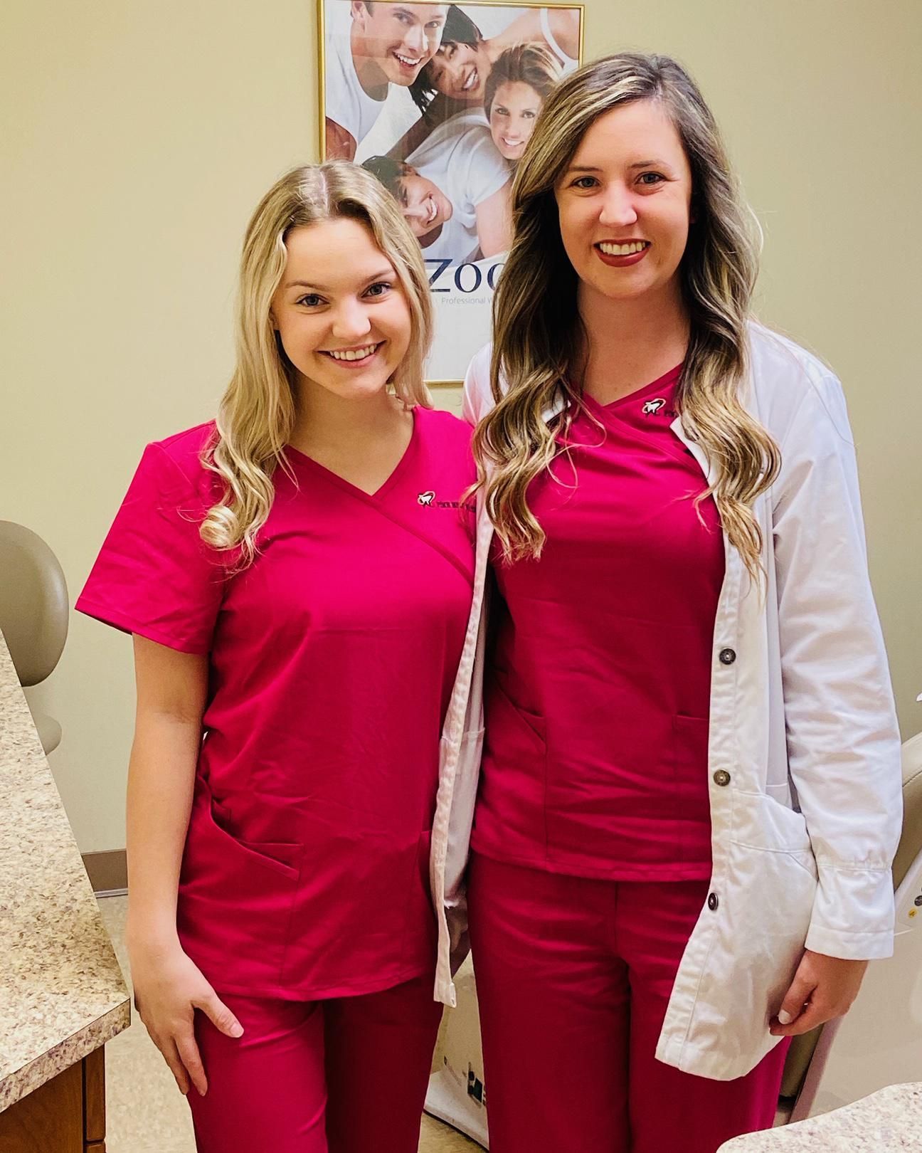 Two dental assistants in pink scrubs pose in an office. One wears a white lab coat.