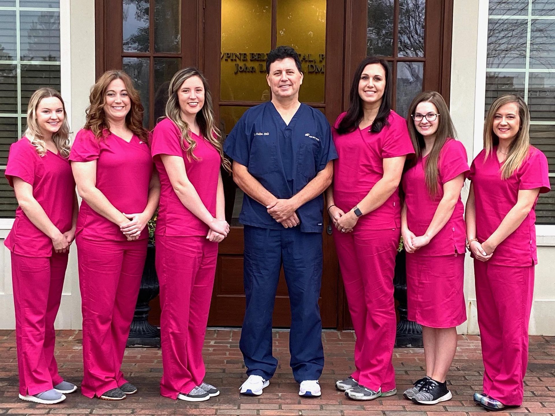 A group of seven people posing in front of a building. Six are wearing pink scrubs, one in blue scrubs.
