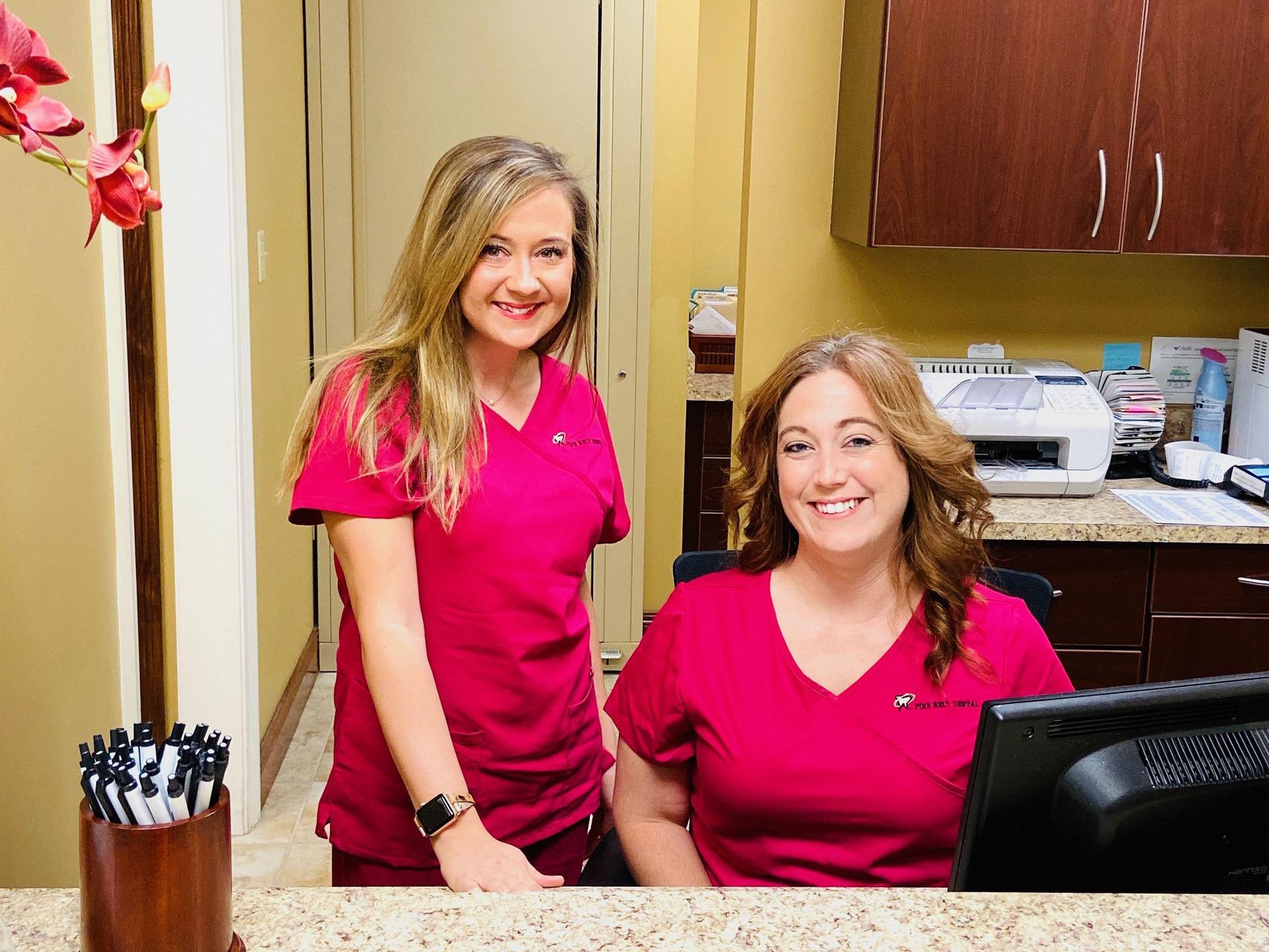 Two women in pink scrubs at a reception desk. One standing, the other sitting, both smiling.