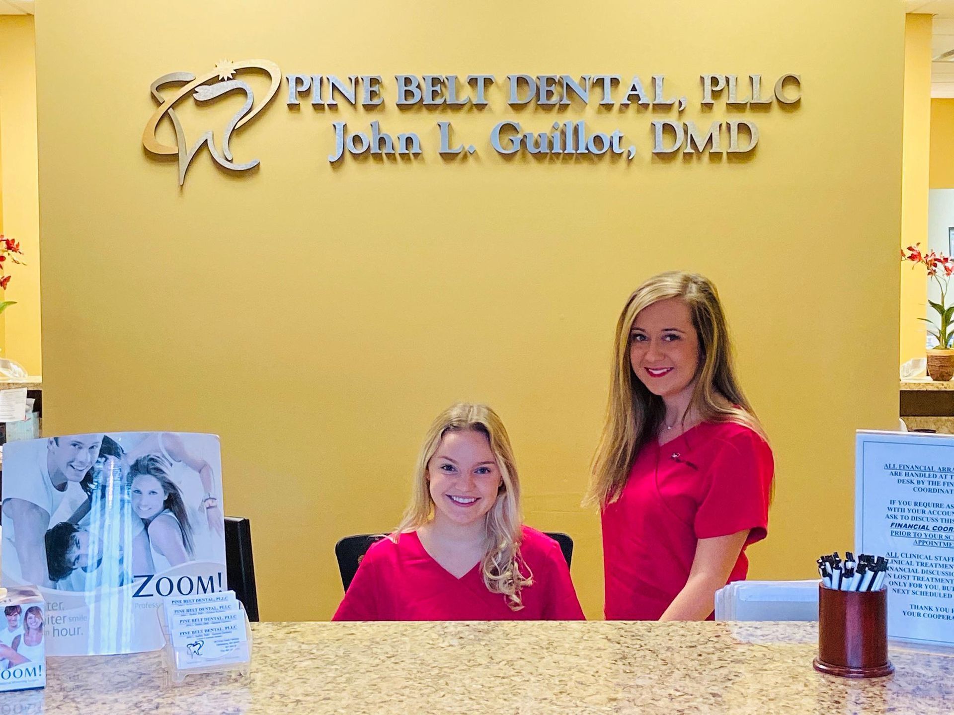 Two women in red shirts behind a reception desk at Pine Belt Dental. Golden wall with logo.