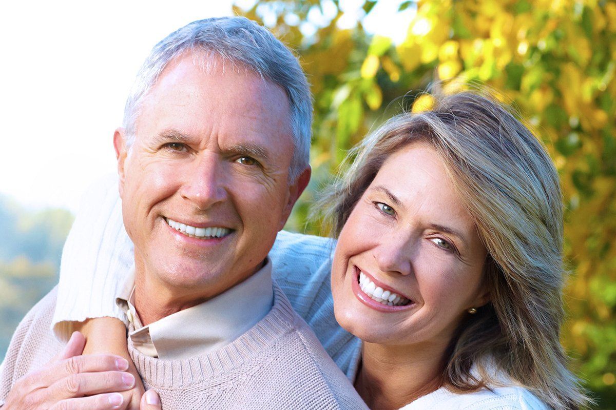 Smiling couple outdoors, man's arms around woman. Light-colored sweaters, blurred green and yellow foliage background.