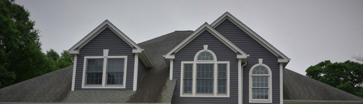 House exterior with gray siding, dormers, and windows under a cloudy sky.