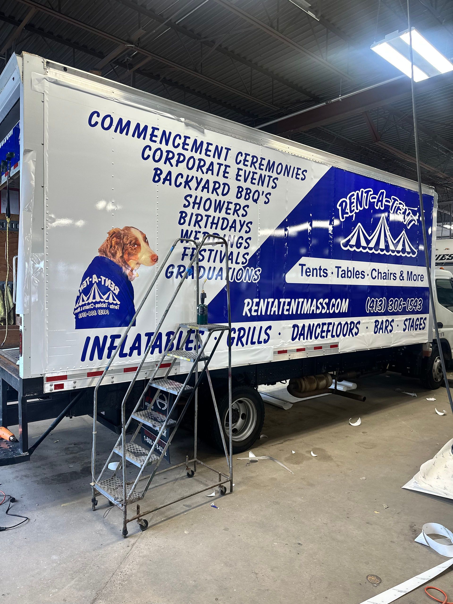 White truck with blue graphics for event rentals, including inflatables and grills. A metal ladder stands near the door.