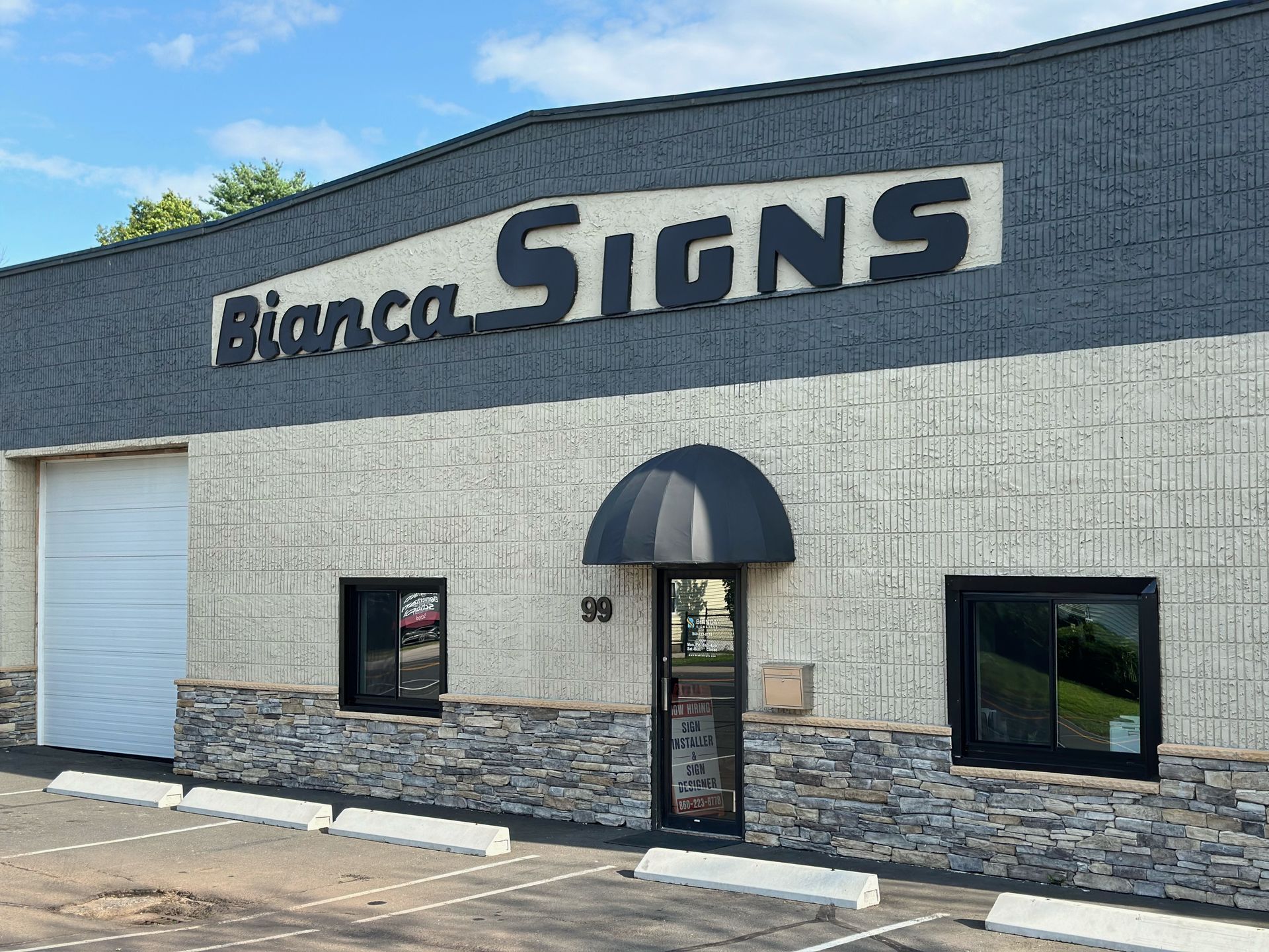 Bianca Signs storefront with black awning, windows, and sign against gray building with stone facade.