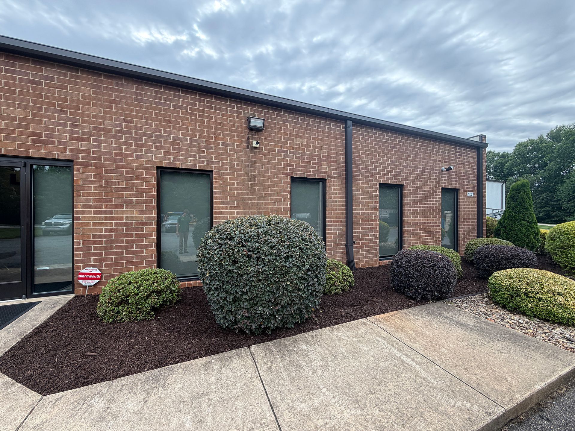 Brick building exterior with tall windows, shrubs, and a concrete walkway under a cloudy sky