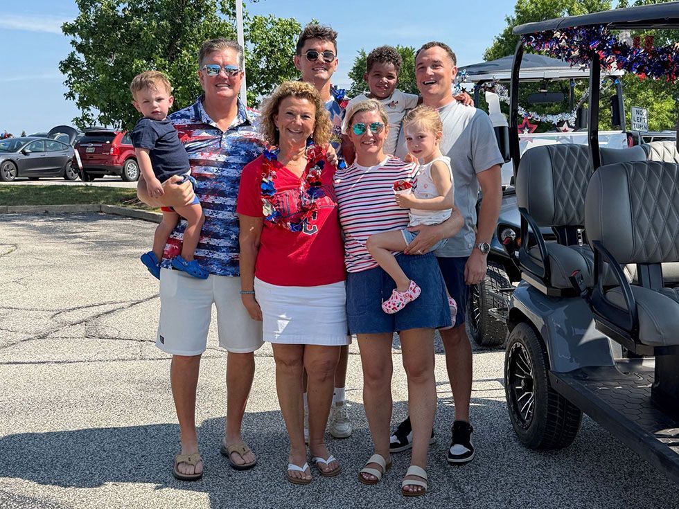 Group of people posing with two toddlers beside a golf cart in a sunny parking lot