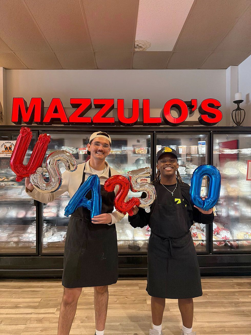 Two employees in black aprons holding red, white, and blue balloon letters