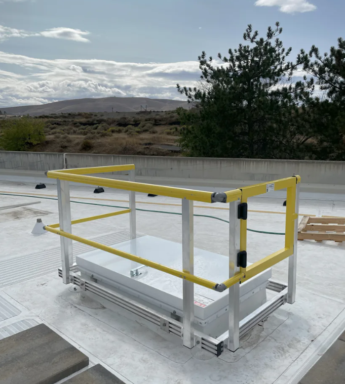 Yellow safety railing installed around a roof access hatch on a flat rooftop against a landscape backdrop.