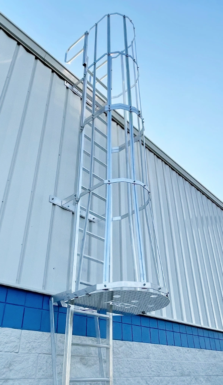 A galvanized steel fixed ladder with a safety cage and a platform mounted to the side of a building against a blue sky.
