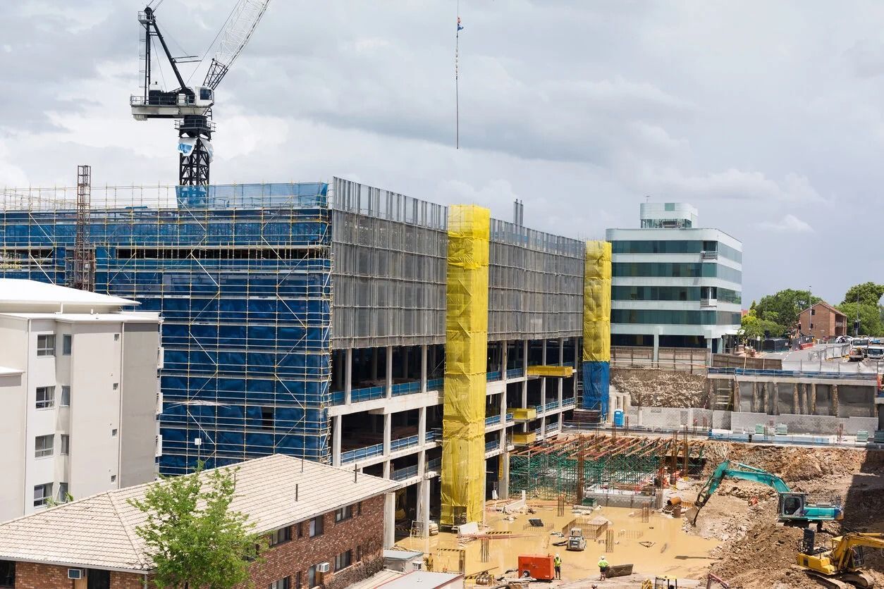 A large construction site featuring a crane, a building wrapped in blue and yellow netting, and nearby excavators.