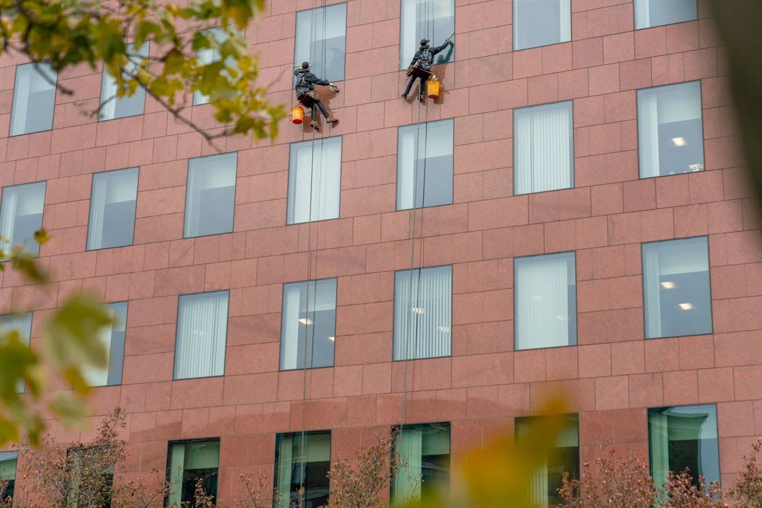 Two workers suspended by ropes clean the windows of a red brick building facade.