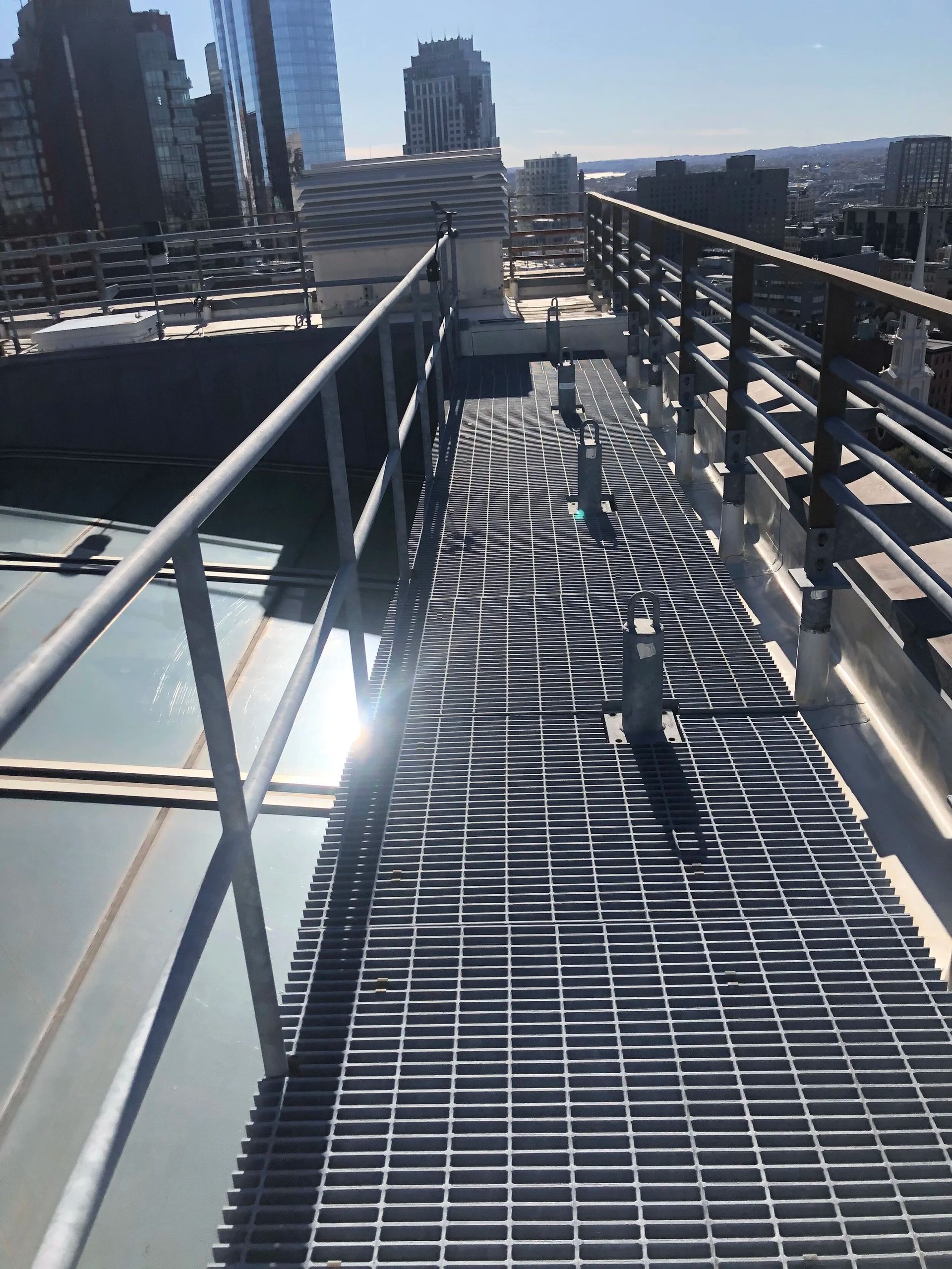 A metal catwalk with railings on a rooftop, overlooking a city skyline on a sunny day.