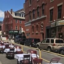 Outdoor dining tables and chairs are set up on a sidewalk beside a street with parked cars and historic red-brick buildings.