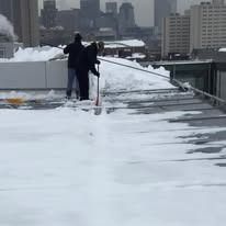 Two people in dark winter gear shovel snow off a flat rooftop against a backdrop of a snowy city skyline.