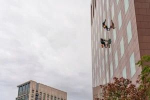 Two people in harnesses rappel down the side of a tall, light-brown building with grid-patterned windows.