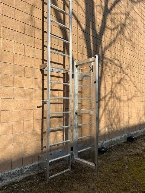 A silver metal ladder with a locked safety gate installed against a tan brick wall.