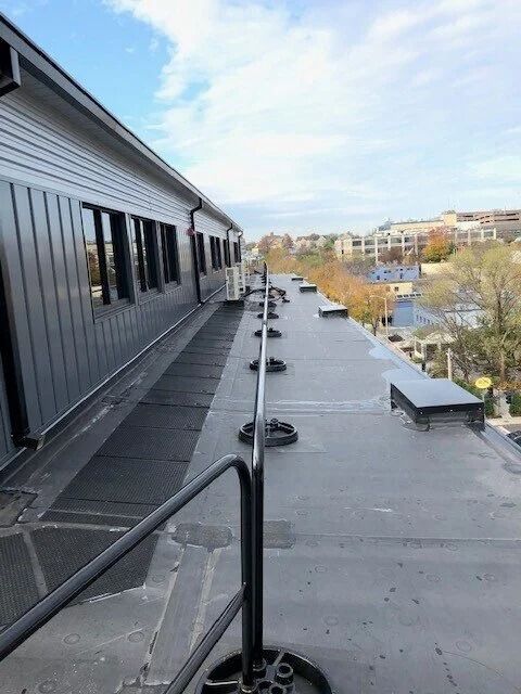 A view along a flat, gray commercial rooftop featuring a safety railing leading to a building with dark-paneled walls.