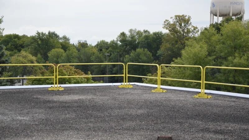 Yellow metal guardrails installed on the edge of a flat, gray gravel roof with trees and a water tower in the background.