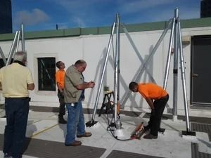 Four workers in a rooftop setting assemble a tall, metal, tripod-like structural support frame.