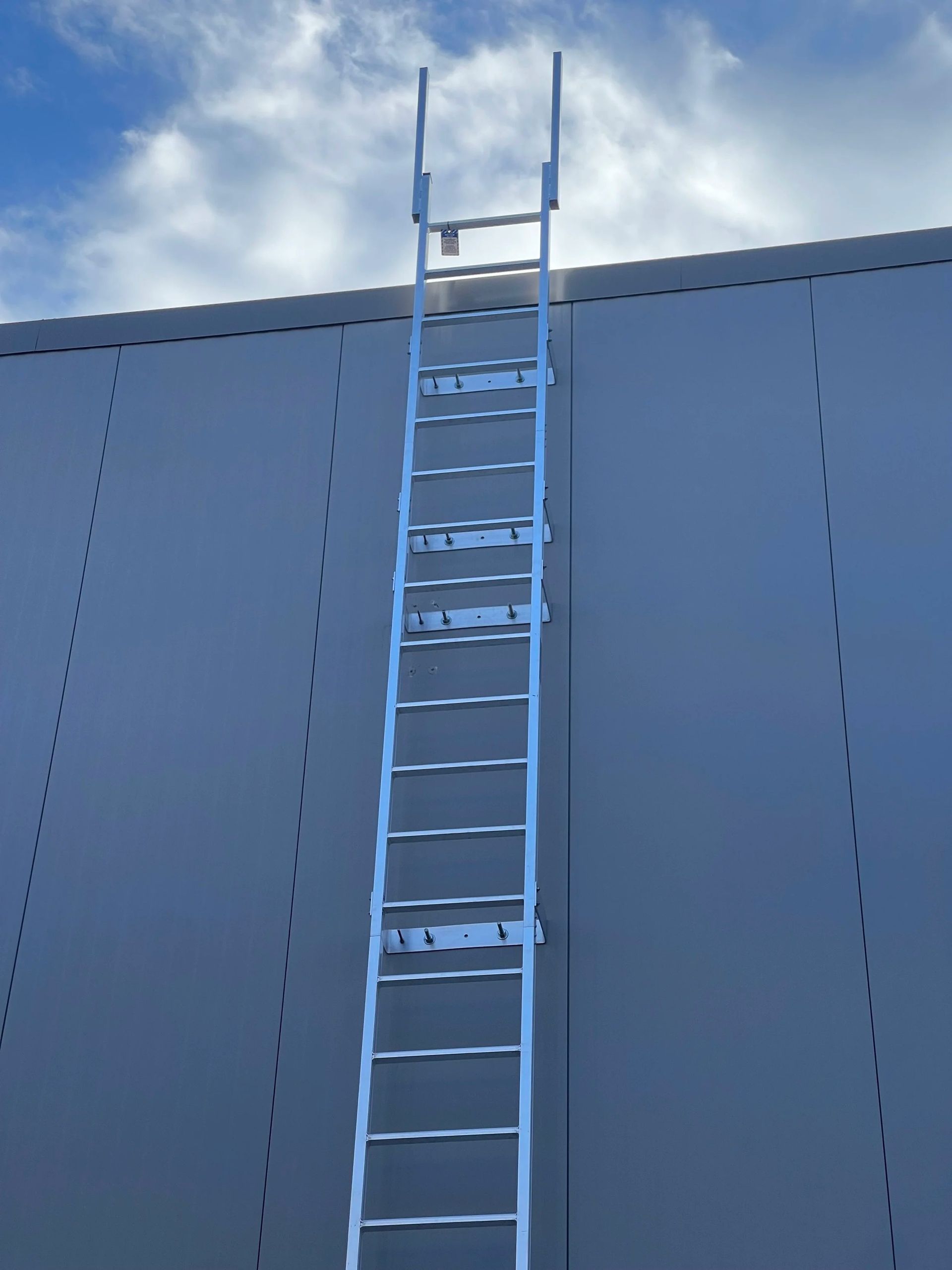 A silver metal ladder extends vertically against a smooth, gray wall under a blue sky with white clouds.