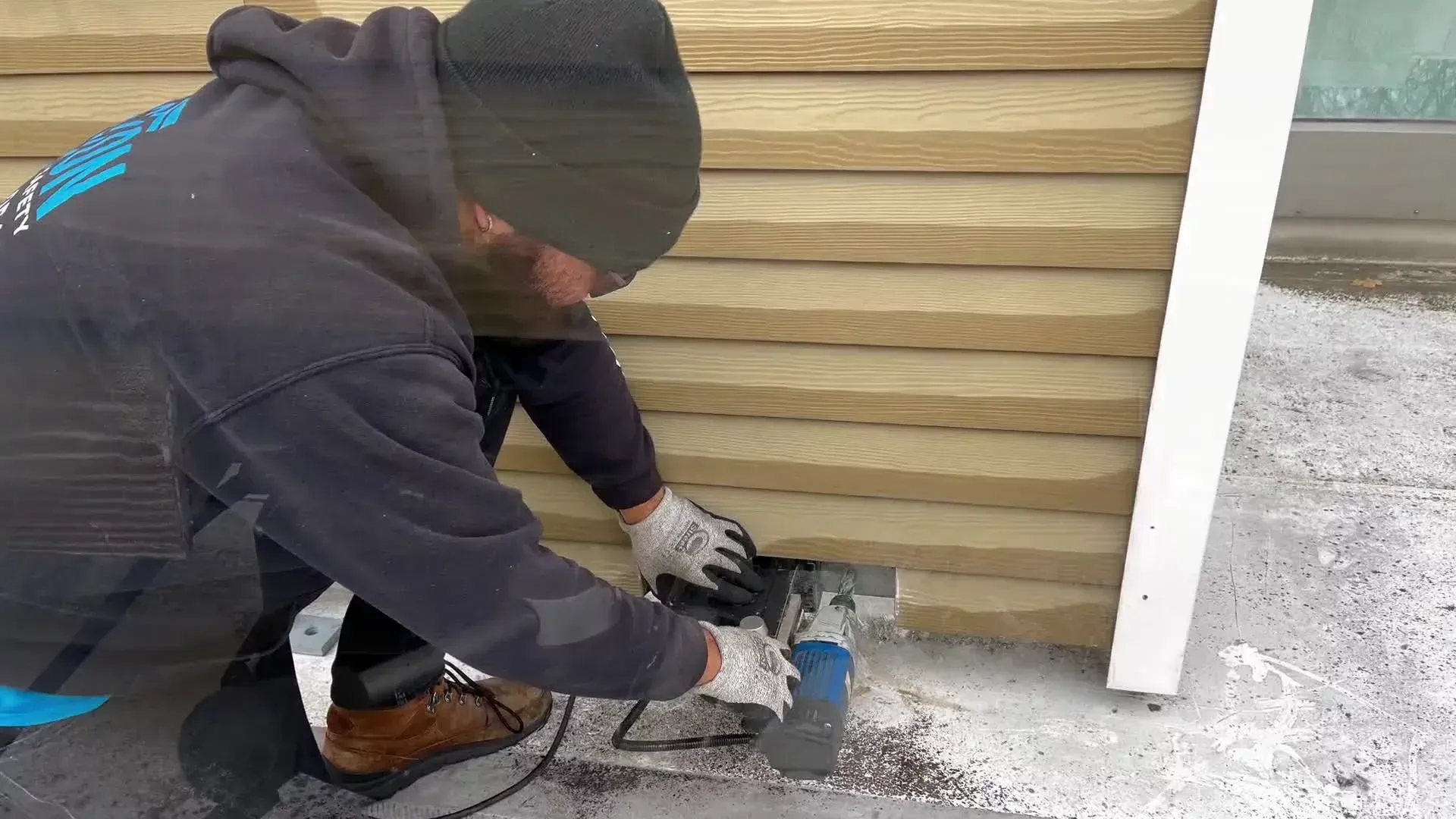 A person in a black hooded sweatshirt and gloves uses a power tool to cut into tan horizontal siding on a building.