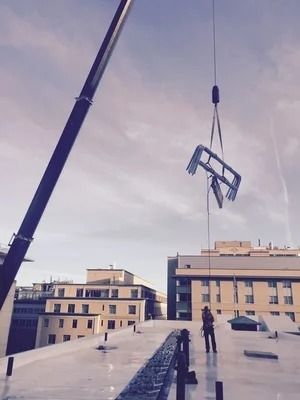 A crane lifts a metal frame above a rooftop where a worker stands under a cloudy sky.