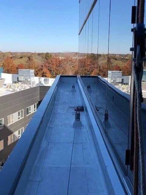 A narrow metal ledge runs alongside a glass building exterior, overlooking trees and nearby rooftops under a blue sky.