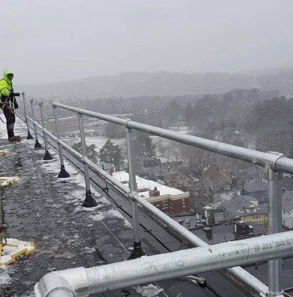 A person in a high-visibility jacket and fall protection gear stands on a snowy rooftop next to a metal safety railing.